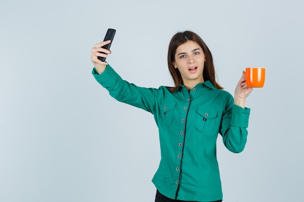 Giovane donna che tiene tazza arancione di tè, prendendo selfie con il cellulare in camicia e guardando fiducioso, vista frontale.