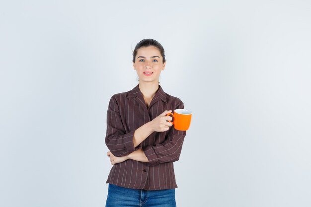 Giovane donna che tiene tazza, guardando la telecamera in camicia, jeans e guardando malinconico, vista frontale.