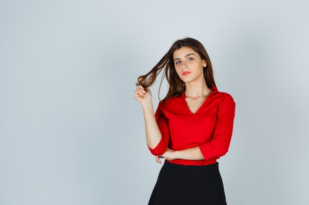 Giovane donna che tiene una ciocca di capelli mentre posa in camicetta rossa, gonna e sembra affascinante