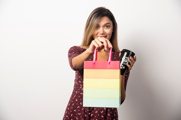 Giovane donna che tiene una piccola borsa del negozio e una tazza di bevanda sul muro bianco.