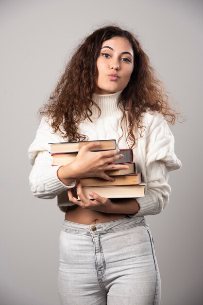 Giovane donna che tiene una pila di libri su un muro grigio. Foto di alta qualità