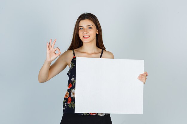 Giovane donna che tiene una tela bianca, che mostra un gesto ok in camicetta, gonna e sembra allegra. vista frontale.