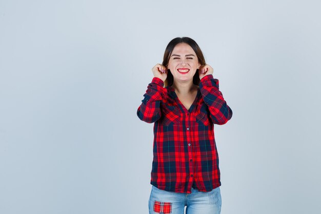 Giovane donna che tira i lobi delle orecchie in camicia a quadri, jeans e sembra carina, vista frontale.