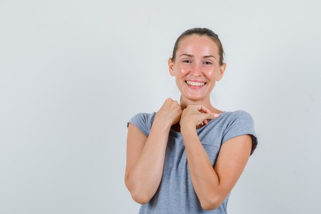 Giovane donna che tocca il colletto in maglietta grigia e che sembra carina. vista frontale.