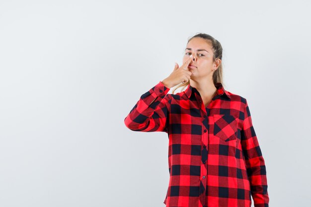 Giovane donna che tocca il naso con il dito in camicia a quadri e sembra carina