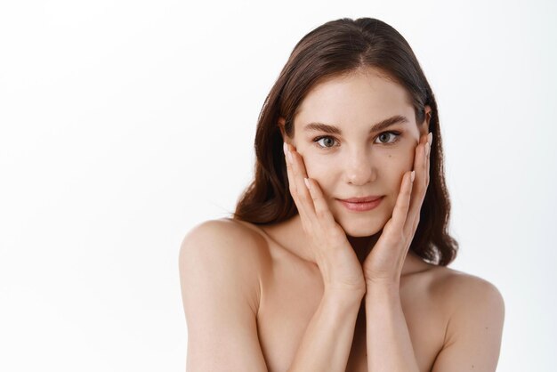 Giovane donna che tocca la pelle del viso idratata pulita e idratata tenendo le mani sulle guance morbide guardando la telecamera in piedi con le spalle nude su sfondo bianco