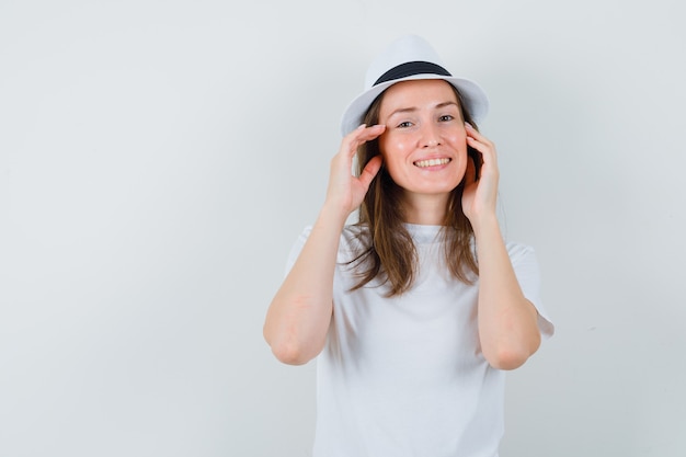 Giovane donna che tocca la pelle del viso sulle guance in maglietta bianca, cappello e un bell'aspetto.