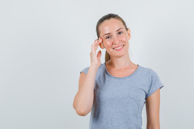 Giovane donna che tocca le tempie con le dita in maglietta grigia e sembra allegra. vista frontale.