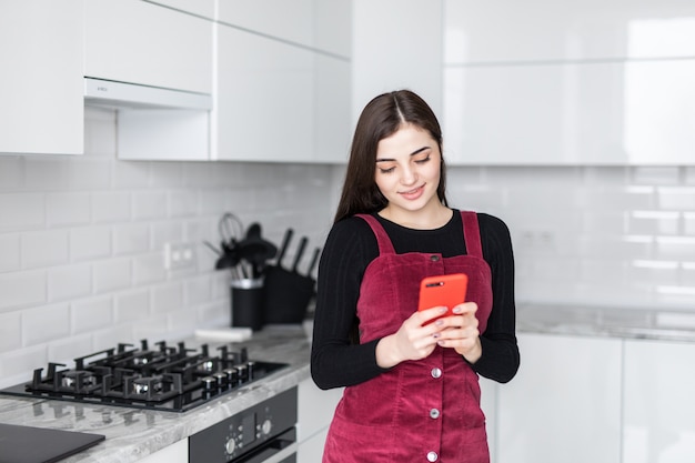 Giovane donna che utilizza smartphone che si appoggia al tavolo da cucina con la tazza da caffè e l'organizzatore in una casa moderna. Messaggio di telefono sorridente della lettura della donna. Ragazza felice del Brunette che scrive un messaggio di testo