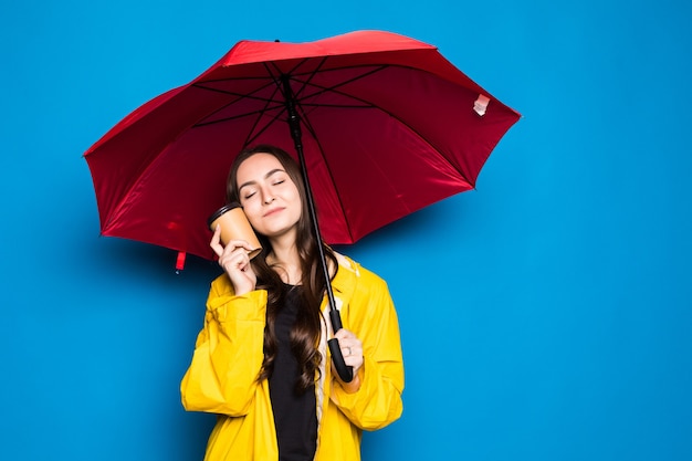 Giovane donna cinese che indossa il cappotto di pioggia che tiene l'ombrello sopra la parete blu isolata molto felice che indica con la mano e il dito