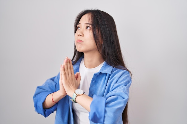 Giovane donna cinese in piedi su sfondo bianco che chiede l'elemosina e prega con le mani insieme all'espressione di speranza sul viso molto emotiva e preoccupata. mendicare.