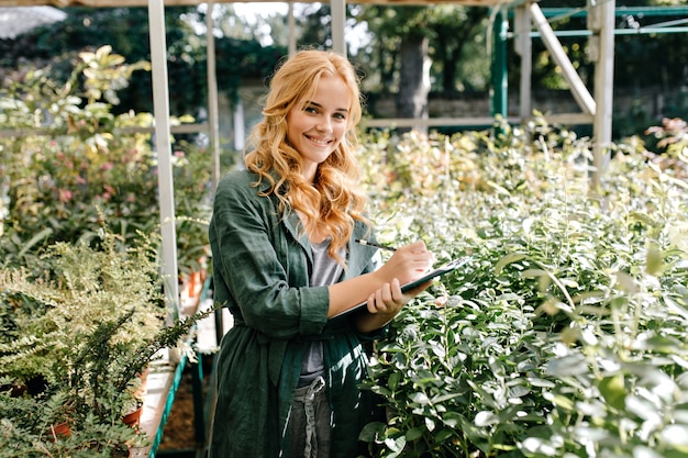 Giovane donna con bei capelli biondi e un sorriso gentile, vestita di una tunica verde con cintura sta lavorando in serra