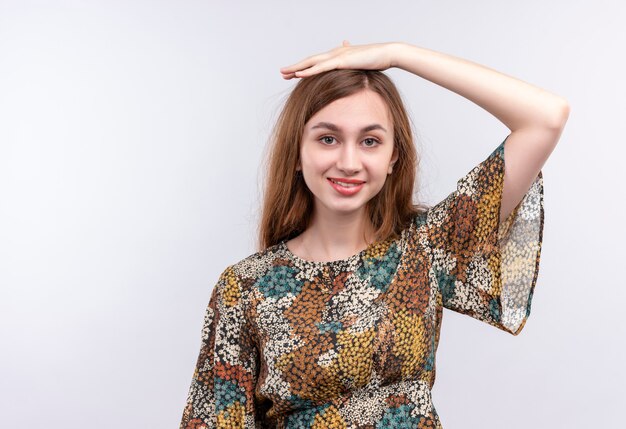 Giovane donna con capelli lunghi che indossa abiti colorati in piedi con la mano sulla sua testa sorridente sul muro bianco