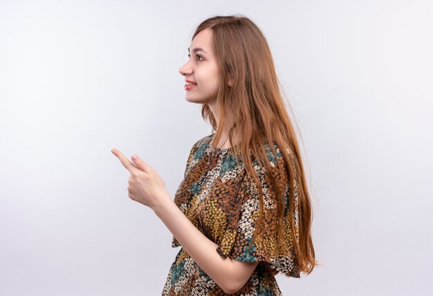 Giovane donna con capelli lunghi che indossa abiti colorati in piedi lateralmente che punta con il dito a lato in piedi sopra il muro bianco