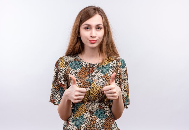 Giovane donna con capelli lunghi che indossa abiti colorati sorridente fiducioso che mostra i pollici in su con entrambe le mani in piedi sul muro bianco