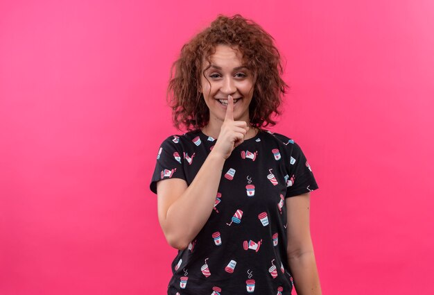 Giovane donna con capelli ricci corti che fa gesto di silenzio con il dito sulle labbra sorridente in piedi sopra il muro rosa