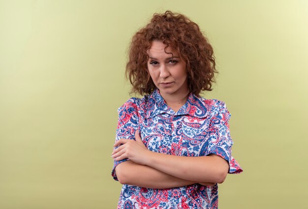 Giovane donna con capelli ricci corti in camicia colorata in piedi con le braccia incrociate sul petto guardando con il viso accigliato oltre il muro verde