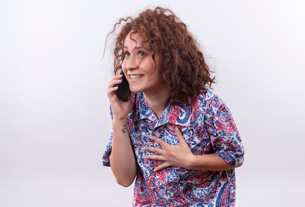 Giovane donna con capelli ricci corti in camicia colorata parlando al telefono cellulare con un timido sorriso sul viso in piedi sopra il muro bianco