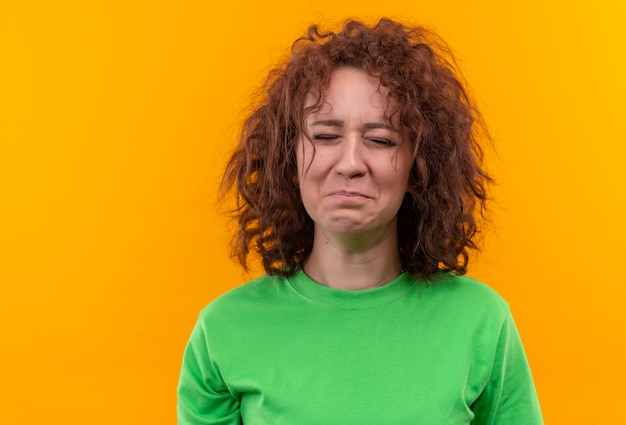 Giovane donna con capelli ricci corti in maglietta verde che piange con la faccia infelice in piedi sopra la parete arancione