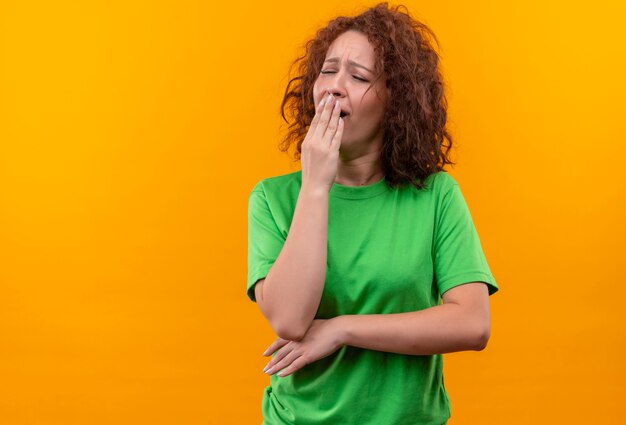 Giovane donna con capelli ricci corti in maglietta verde che sbadiglia che copre la bocca con la mano in piedi sopra la parete arancione