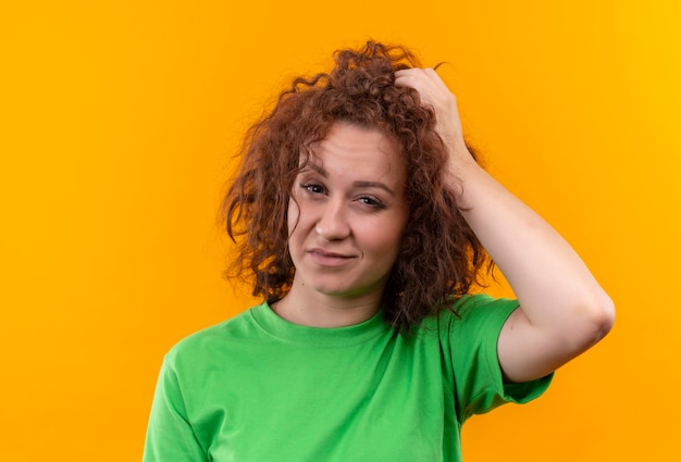 Giovane donna con capelli ricci corti in maglietta verde che sembra confusa e molto ansiosa di toccare la sua testa in piedi