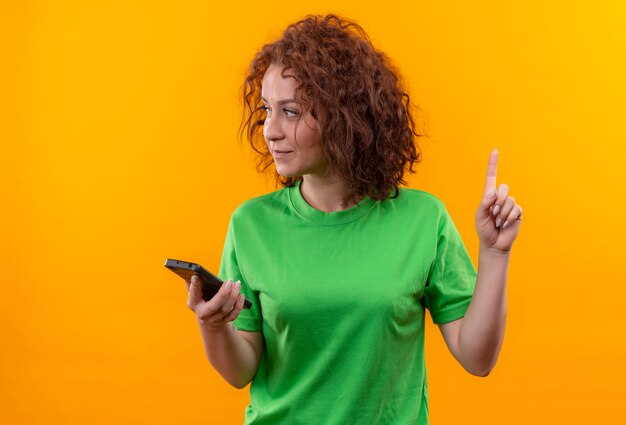 Giovane donna con capelli ricci corti in maglietta verde che tiene smartphone puntare il dito verso l'alto con la faccia seria in piedi