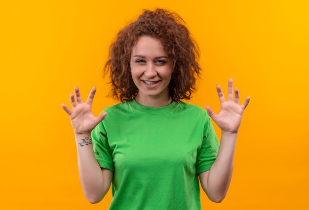 Giovane donna con capelli ricci corti in maglietta verde sorridente che fa gesto di artigli di gatto in piedi sopra la parete arancione