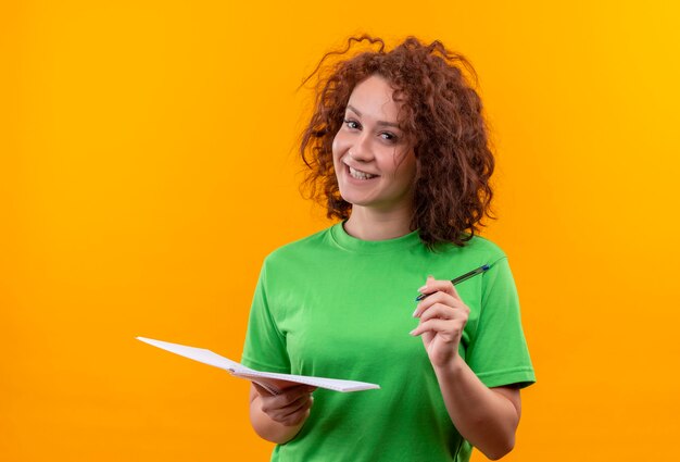 Giovane donna con capelli ricci corti in t-shirt verde che tiene taccuino e penna sorridente in piedi positivo e felice