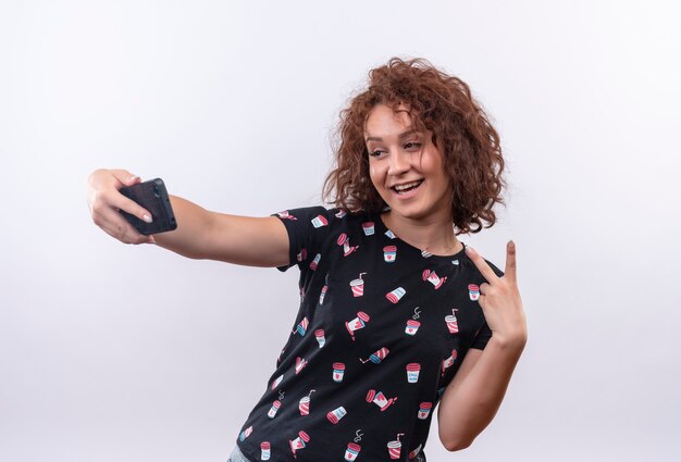 Giovane donna con capelli ricci corti prendendo selfie utilizzando il suo smartphone sorridendo alla telecamera che mostra il segno di vittoria in piedi sul muro bianco