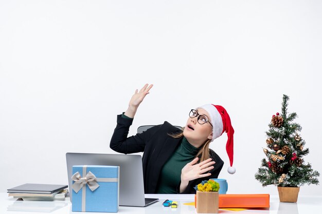 Giovane donna con cappello di Babbo Natale seduto a un tavolo con un albero di Natale e un regalo