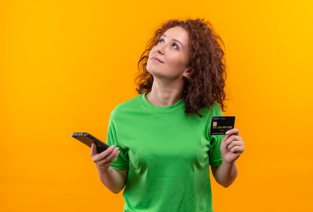 Giovane donna con corti capelli ricci in maglietta verde che tiene due smartphone guardando con espressione pensierosa sul viso cercando di fare la scelta in piedi