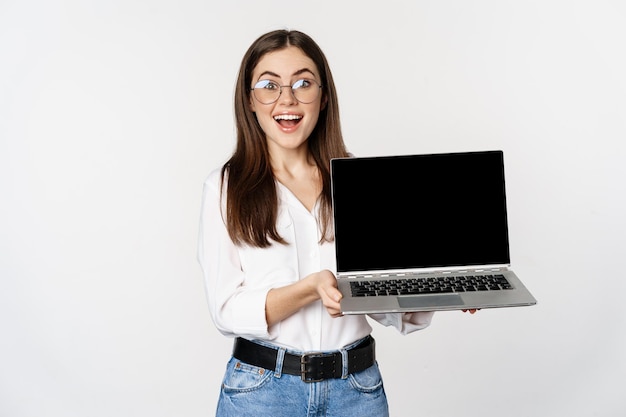 Giovane donna con gli occhiali che mostra lo schermo del laptop, dimostrando una promozione su computer, sito Web o negozio, in piedi su sfondo bianco.