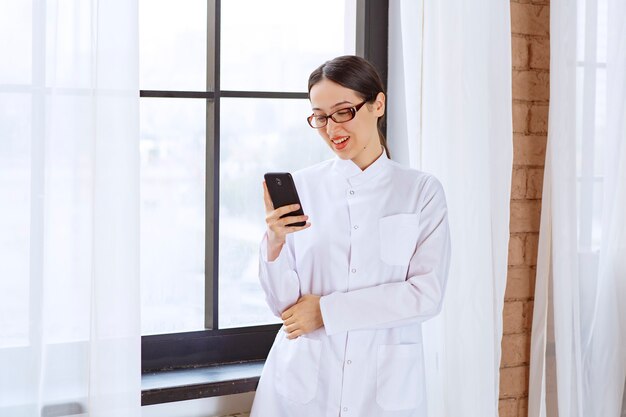 Giovane donna con gli occhiali in camice da laboratorio che tiene i messaggi di controllo sul cellulare vicino alla finestra.