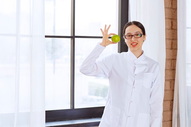 Giovane donna con gli occhiali in camice da laboratorio che tiene mela verde vicino alla finestra.