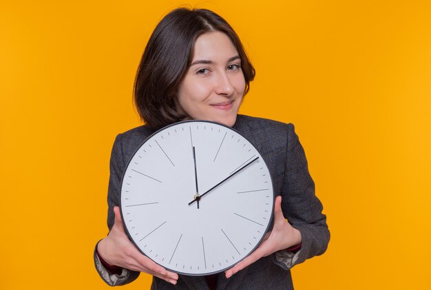 Giovane donna con i capelli corti che indossa giacca grigia che tiene orologio da parete guardando davanti sorridendo allegramente felice e positivo in piedi sopra la parete arancione