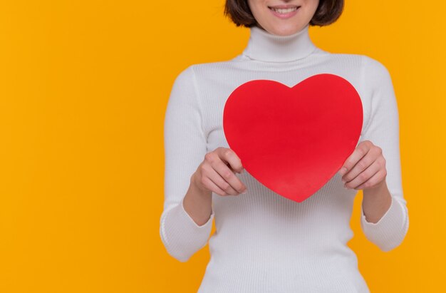 Giovane donna con i capelli corti che tiene il cuore fatto di cartone che sorride allegramente in piedi sopra la parete arancione