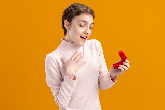 giovane donna con i capelli corti che tiene scatola rossa con anello di fidanzamento guardandolo stupito e felice concetto di San Valentino in piedi sul muro arancione