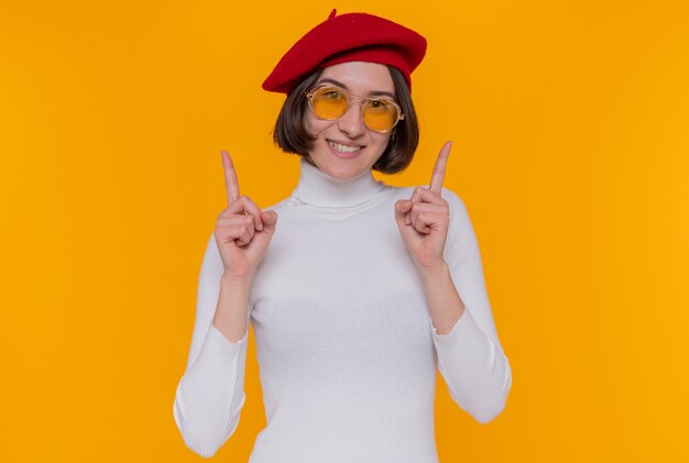 Giovane donna con i capelli corti in dolcevita bianco che indossa berretto e occhiali da sole gialli guardando davanti felice e positivo che mostra le dita indice sorridendo allegramente in piedi sopra la parete arancione