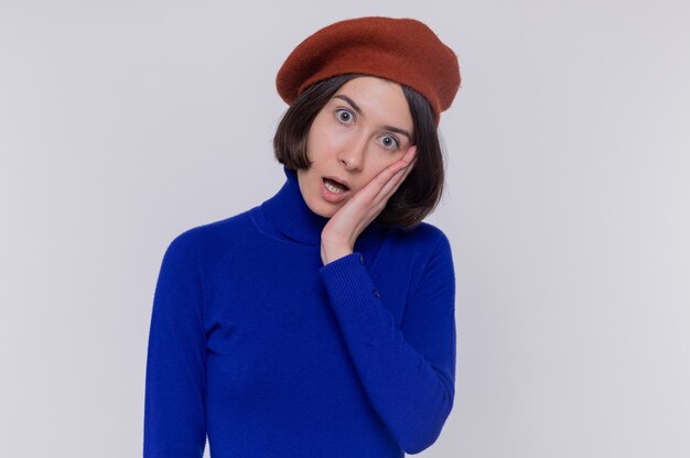 Giovane donna con i capelli corti in dolcevita blu che indossa berretto guardando davanti confuso e sorpreso in piedi sopra il muro bianco