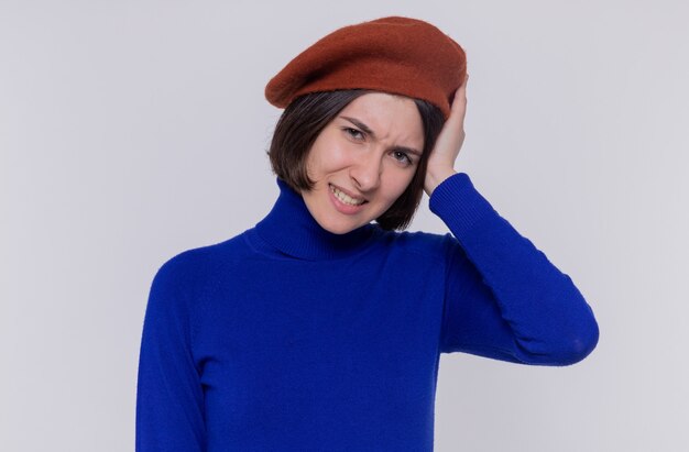 Giovane donna con i capelli corti in dolcevita blu che indossa berretto guardando davanti confuso tenendo la testa sulla sua testa per errore tanding sul muro bianco