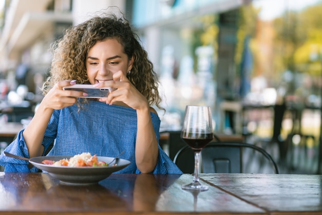 Giovane donna con i capelli ricci che scatta foto del suo cibo con un telefono cellulare mentre pranza in un ristorante.