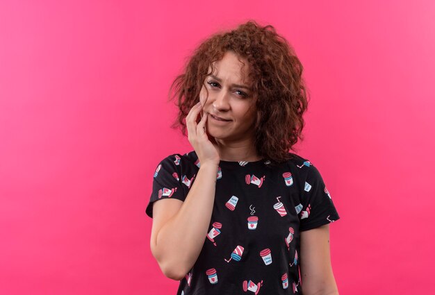 Giovane donna con i capelli ricci corti cercando toccando la guancia sensazione di dolore in piedi sopra la parete rosa