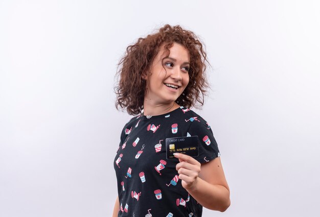 Giovane donna con i capelli ricci corti che mostra la carta di credito che osserva da parte sorridente che sta sopra la parete bianca