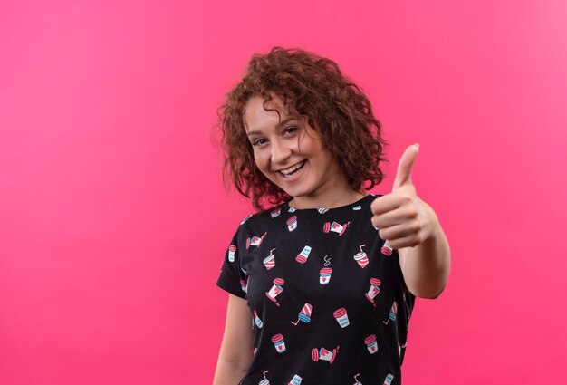 Giovane donna con i capelli ricci corti che sorride allegramente mostrando i pollici in su in piedi sopra il muro rosa