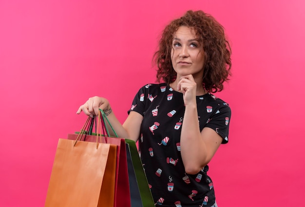 Giovane donna con i capelli ricci corti che tiene i sacchetti di carta che osserva in su con l'espressione pensierosa sul fronte che prova a fare la scelta che sta sopra la parete rosa