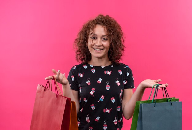 Giovane donna con i capelli ricci corti che tiene i sacchetti di carta sorridendo allegramente felice e positivo in piedi sopra il muro rosa