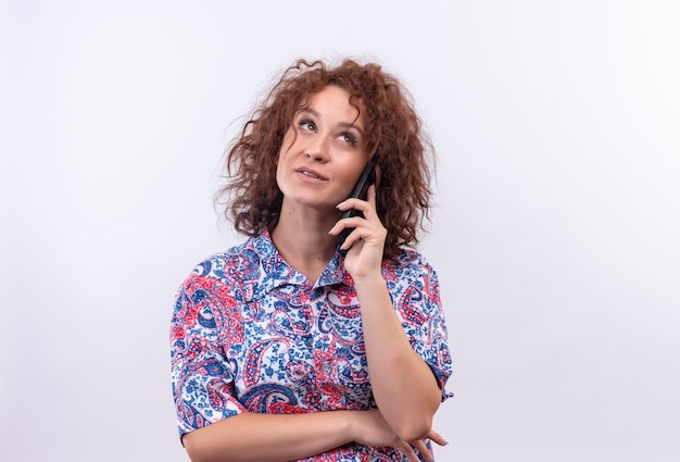 Giovane donna con i capelli ricci corti in camicia colorata alzando lo sguardo pensando mentre parla al telefono cellulare sopra il muro bianco