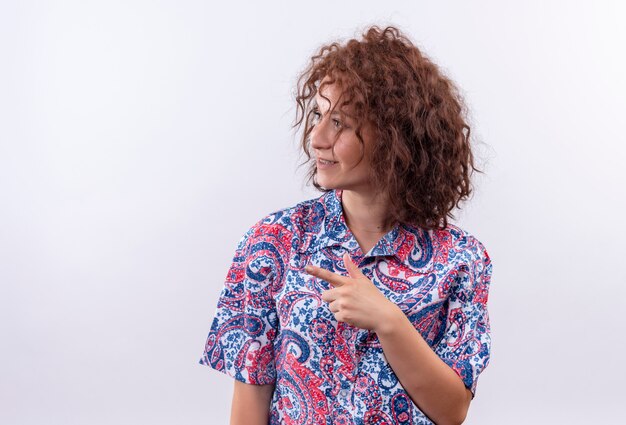 Giovane donna con i capelli ricci corti in camicia colorata che osserva da parte sorridente che punta con il dito di lato sopra il muro bianco