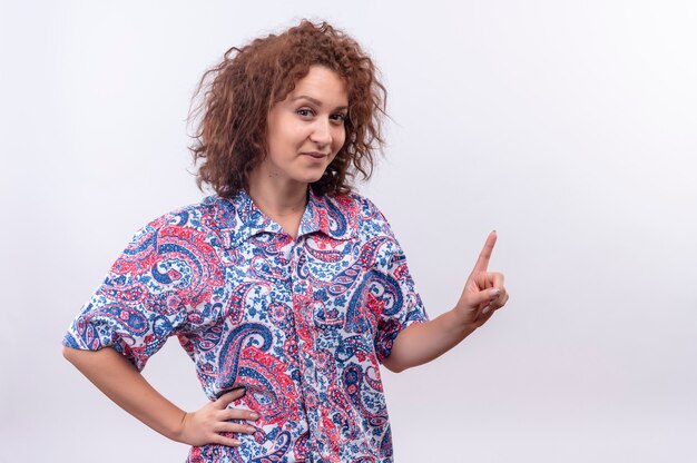 Giovane donna con i capelli ricci corti in camicia colorata felice e positivo guardando e rivolto verso l'alto con il dito in piedi sul muro bianco