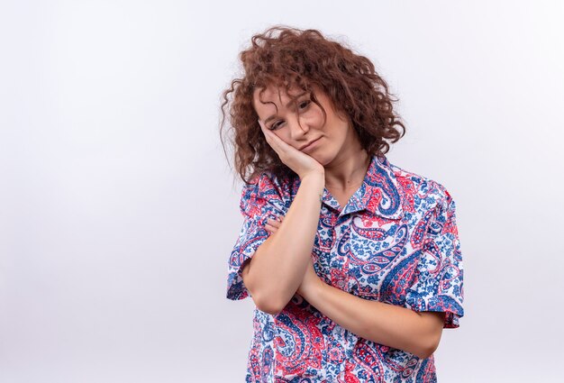 Giovane donna con i capelli ricci corti in camicia colorata guardando la testa pendente sul suo braccio stanco e annoiato in piedi sul muro bianco